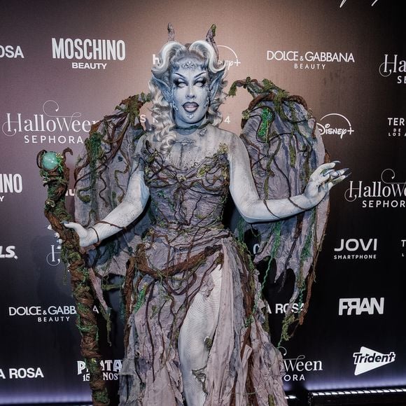 Gloria Groove veste uma túnica escultural em tons cinzentos e terrosos com acabamento desgastado e aplicação de ramos e raízes secas por todo o traje, simulando uma criatura da floresta ou gárgula. A caracterização inclui asas em forma de teia/raízes nas costas, pintura corporal e facial acinzentada, chifres prateados na cabeça e um cajado ornamentado com uma pedra/esfera verde