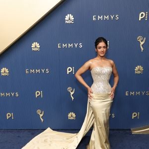 Shabana Azeez veste um modelo tomara que caia de dois tons, composto por corset prateado totalmente bordado com pedrarias e padrões geométricos. A saia reta em cetim champanhe traz um drapeado sobreposto no quadril que se estende para uma cauda lateral longa e fluida. O visual é arrematado por um penteado preso sofisticado