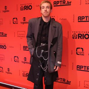 João Cortes aposta em uma composição ousada e desconstruída de estilo grunge. Ele veste uma camisa social preta sob um paletó oversized cinza de abotoamento duplo com lapelas recortadas em preto, combinado a uma calça escura de alfaiataria sobreposta por uma espécie de saia utilitária ou aba longa com ilhoses. O visual é complementado por correntes metálicas, alfinetes de segurança no cós e sapatos pretos tratorados pesados