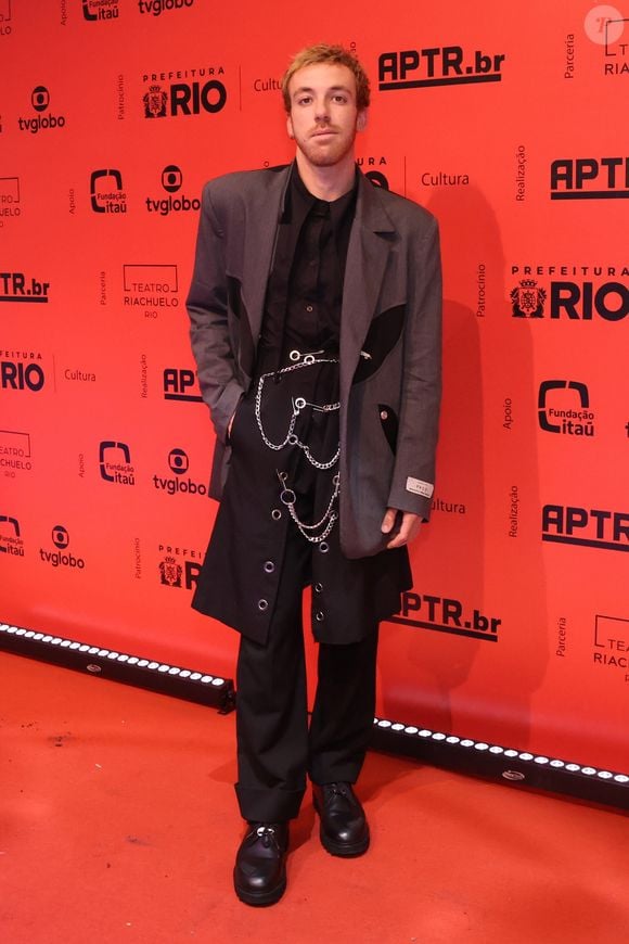 João Cortes aposta em uma composição ousada e desconstruída de estilo grunge. Ele veste uma camisa social preta sob um paletó oversized cinza de abotoamento duplo com lapelas recortadas em preto, combinado a uma calça escura de alfaiataria sobreposta por uma espécie de saia utilitária ou aba longa com ilhoses. O visual é complementado por correntes metálicas, alfinetes de segurança no cós e sapatos pretos tratorados pesados