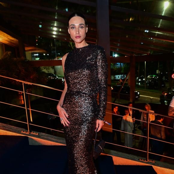 Débora Nascimento: a atriz usa um vestido longo tubinho de manga única e um ombro só, inteiramente bordado em paetês escuros de tom acobre e preto. O visual traz cabelos presos em um coque polido, unhas escuras, joias discretas e uma pequena bolsa preta de mão