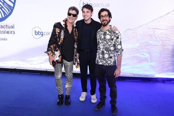 Jorge Israel, Dani Flomin e Leo Israel: Jorge (à esquerda) combina blazer estampado com tigres, calça de padronagem geométrica psicodélica em preto e branco e óculos escuros. Dani (ao centro) usa terno azul-escuro minimalista com camiseta e tênis branco. Leo (à direita) veste camisa de botões com estampa de círculos preto e branco e calça jeans escura