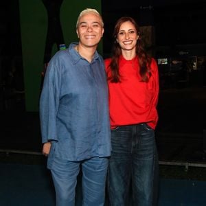 A cantora Maria Gadú surge com um conjunto monocromático de linho azul-serenity, composto por camisa de botões com mangas longas e modelagem solta, acompanhada por calça de corte reto no mesmo tecido, além de tênis brancos de cano baixo. Ao seu lado, Ana Luiza veste uma blusa vermelha vibrante de gola alta discreta combinada com calça jeans wide leg escura e tênis bicolores em vermelho e branco