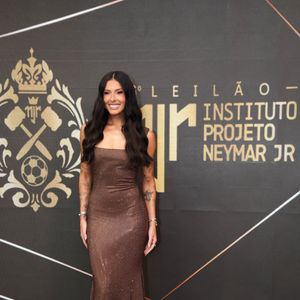 Ana Castela escolheu um vestido longo bronze com acabamento cintilante e brilho sutil, de modelagem reta, decote de ombro a ombro com alças finas e caimento rente ao corpo
