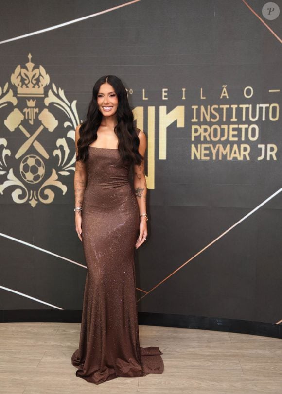 Ana Castela escolheu um vestido longo bronze com acabamento cintilante e brilho sutil, de modelagem reta, decote de ombro a ombro com alças finas e caimento rente ao corpo