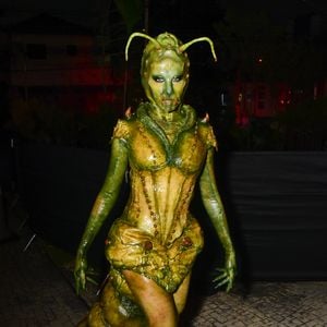 Camilla Pudim usa fantasia de inseto escultural e texturizada em tons de verde e amarelo-esverdeado. O figurino parece uma armadura de corpo inteiro com aspecto orgânico e brilhante, apresentando costuras proeminentes em forma de contas que descem pelo corpete ajustado