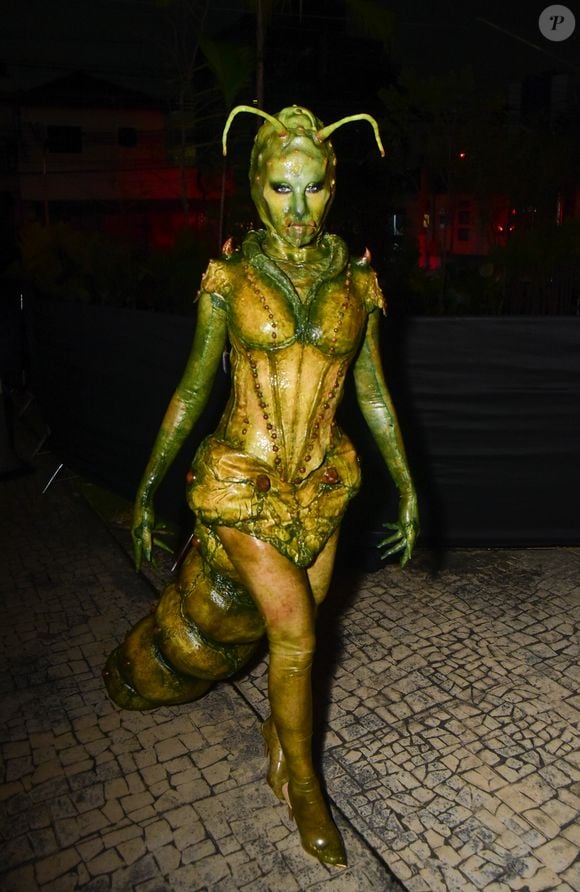 Camilla Pudim usa fantasia de inseto escultural e texturizada em tons de verde e amarelo-esverdeado. O figurino parece uma armadura de corpo inteiro com aspecto orgânico e brilhante, apresentando costuras proeminentes em forma de contas que descem pelo corpete ajustado