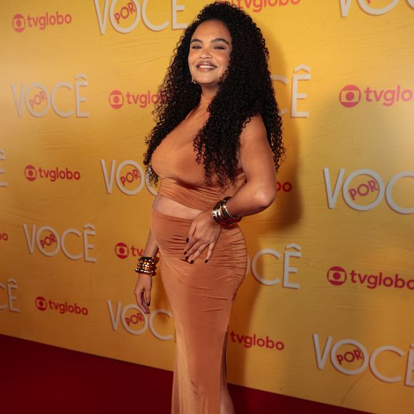 Lucy Ramos destaca a gestação com sofisticação em um conjunto de duas peças em tom caramelo, composto por crop top e saia longa drapeada com fenda lateral. O look ganha impacto com múltiplos braceletes dourados nos dois pulsos e sandálias de tiras finas tom sobre tom