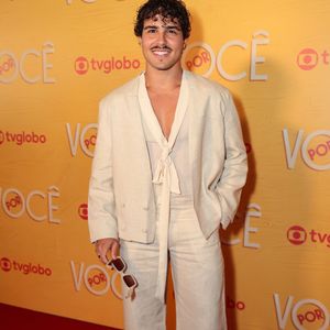 Igor Jansen traz um conjunto monocromático off-white com blazer curto de abotoamento duplo, calça wide leg de cintura alta e blusa fluida com amarração em laço no decote. O artista segura óculos de sol claros nas mãos e completa o look com sapatos em tom neutro