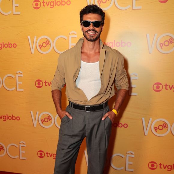 Ator combina estilo clássico e descontraído usando camisa cáqui aberta sobre regata branca básica, usada por dentro da calça cinza de alfaiataria com pregas e cinto preto. A produção ganha atitude com óculos escuros estilo wayfarer e sapatos pretos de sola robusta
