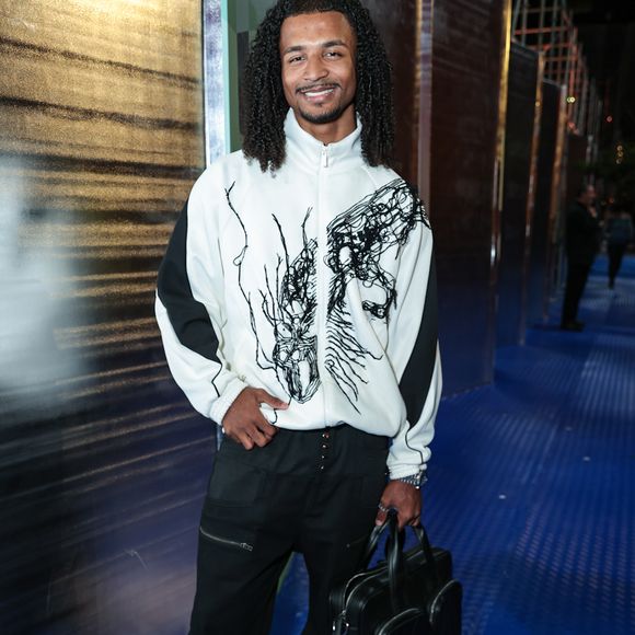 O influenciador Matheus Tet trouxe uma abordagem esportiva de luxo, vestindo uma jaqueta tracksuit branca com grafismos em preto que lembram traços de nanquim orgânicos. A peça de gola alta foi combinada a calças pretas de estilo cargo com detalhes de zíperes horizontais, unindo funcionalidade e design