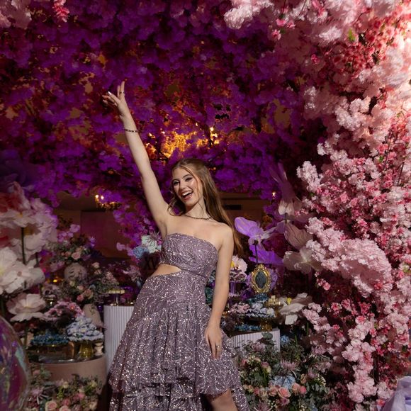 Esse é o tipo de vestido que transforma a debutante em estrela de editorial. O tecido com leve efeito acetinado refletia a iluminação rosa da decoração floral, criando um jogo de cores absolutamente instagramável. É dramático, é moderno e tem aquela pegada de alta-costura jovem.