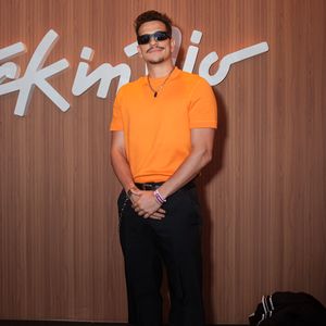 Victor Lamoglia aposta em um visual smart casual vibrante e moderno. O ator veste uma camisa polo em tom laranja vivo combinada com calça de alfaiataria preta de corte reto e cinto com correntes metálicas pendentes, arrematando o look com botas pretas e óculos escuros