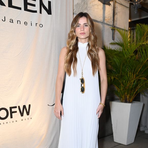 Giulia Be apostou em um minimalismo etéreo que flerta com o neoclássico. Ela veste um vestido longo off-white inteiramente plissado, com gola alta e modelagem sem mangas que valoriza a fluidez do tecido sob a brisa carioca