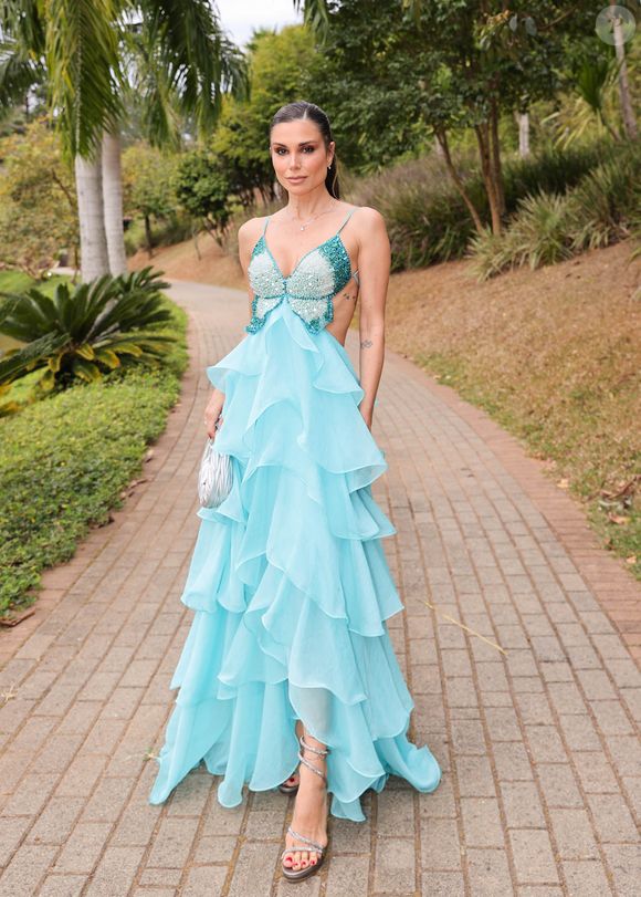 Flávia Viana apostou em um look sofisticado para o casamento, com um vestido longo em tom azul turquesa que transmite frescor e leveza. A peça tem o busto todo bordado em pedrarias reluzentes que formam o desenho de uma borboleta, enquanto as laterais vazadas realçam a silhueta. A saia, confeccionada em camadas fluidas de organza, cria um efeito esvoaçante e volumoso, conferindo movimento a cada passo