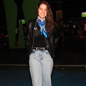 A atriz Nathalia Dill opta pelo contraste urbano combinando uma calça jeans reta de lavagem clara e cintura alta com uma jaqueta de couro preta em estilo biker. Por baixo da jaqueta, veste uma blusa preta sóbria, que ganha um ponto de cor marcante com um lenço de seda amarrado ao pescoço em tons de azul e branco, finalizando com botas pretas de amarrar