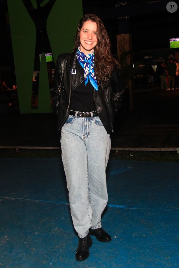 A atriz Nathalia Dill opta pelo contraste urbano combinando uma calça jeans reta de lavagem clara e cintura alta com uma jaqueta de couro preta em estilo biker. Por baixo da jaqueta, veste uma blusa preta sóbria, que ganha um ponto de cor marcante com um lenço de seda amarrado ao pescoço em tons de azul e branco, finalizando com botas pretas de amarrar