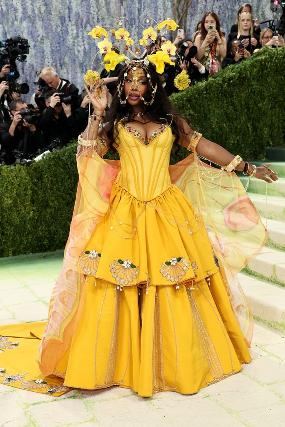 SZA: a cantora apresentou uma explosão de cor com um vestido amarelo vibrante inspirado na flora tropical. A peça contou com um espartilho estruturado e uma saia em camadas decorada com conchas bordadas, mas o grande destaque foi o adereço de cabeça: uma estrutura de arame dourado que suspendia orquídeas amarelas e detalhes em ouro, emoldurando seu rosto com uma delicadeza luxuosa