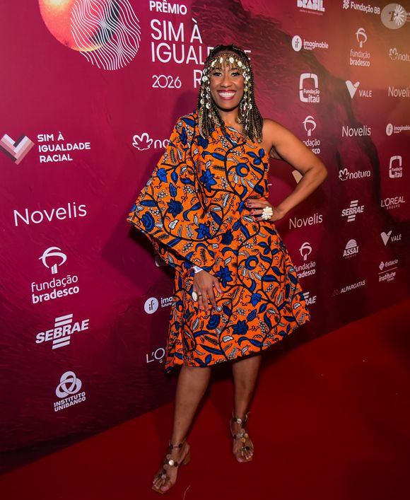 Na imagem Luana Xavier, a artista escolheu um vestido curto de um ombro só com uma modelagem ampla e estruturada, destacando-se pela estampa vibrante em tons de laranja, azul e detalhes florais. O visual ganha um peso cultural marcante com o uso de um acessório de cabeça elaborado com búzios e elementos metálicos, que emoldura o rosto de forma imponente