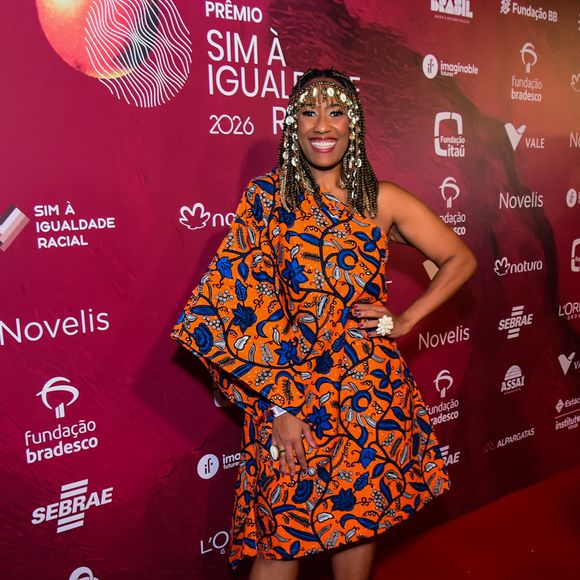 Na imagem Luana Xavier, a artista escolheu um vestido curto de um ombro só com uma modelagem ampla e estruturada, destacando-se pela estampa vibrante em tons de laranja, azul e detalhes florais. O visual ganha um peso cultural marcante com o uso de um acessório de cabeça elaborado com búzios e elementos metálicos, que emoldura o rosto de forma imponente