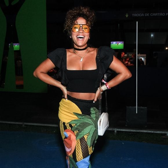 A famosa exibe um visual tropical com saia lápis de comprimento midi com fenda lateral sutil e uma vibrante estampa de arara e folhagens sobre fundo preto e amarelo. Na parte superior, optou por um cropped preto de mangas bufantes estruturadas e decote quadrado, complementado por óculos de lentes amarelas translúcidas, gargantilha preta e sandálias brancas de salto