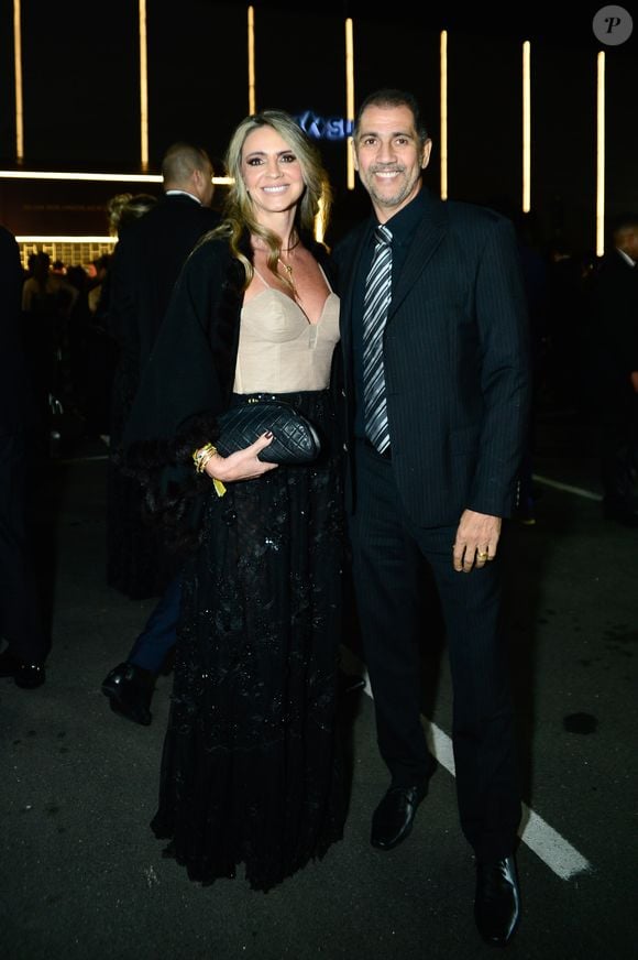 Ex jogador Giovanni e a esposa: o ex-goleiro vestiu um terno escuro risca-de-giz com gravata prateada padronizada, enquanto sua acompanhante optou por um vestido longo preto de saia fluida com detalhes cintilantes e top estruturado estilo corset em tom bege/nude, além de uma capa preta sobreposta