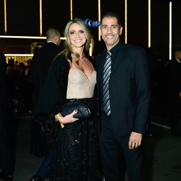 Ex jogador Giovanni e a esposa: o ex-goleiro vestiu um terno escuro risca-de-giz com gravata prateada padronizada, enquanto sua acompanhante optou por um vestido longo preto de saia fluida com detalhes cintilantes e top estruturado estilo corset em tom bege/nude, além de uma capa preta sobreposta