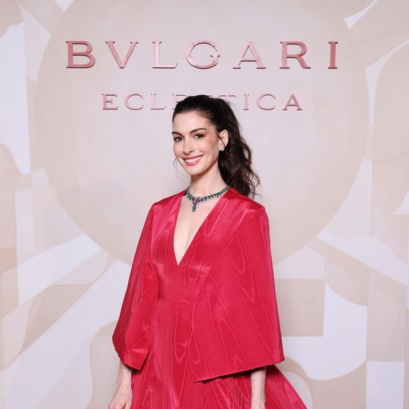 Anne Hathaway apostou em um vestido volumoso de alta-costura da Valentino, confeccionado em seda vermelha moiré, com decote profundo e uma capa elegante que caía sobre os ombros e costas