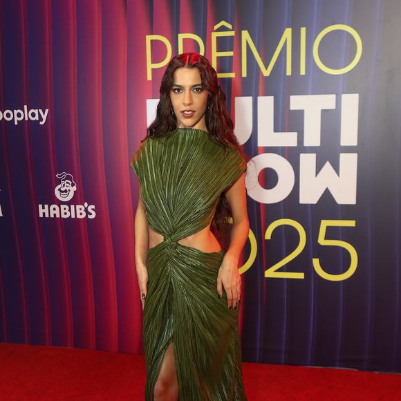 Marina Sena surgiu com um vestido verde de efeito plissado que combina escultura e fluidez de maneira precisa. A peça apresenta um trabalho têxtil que imita folhas texturizadas, formando pregas alongadas que desenham o corpo em direção ao centro, onde um torcido frontal cria um recorte estratégico e deixa a cintura à mostra. O decote estruturado sobe como uma gola aberta, moldando o colo com um volume arquitetônico que contrasta com a fenda frontal profunda, responsável por alongar a silhueta e revelar o movimento da saia