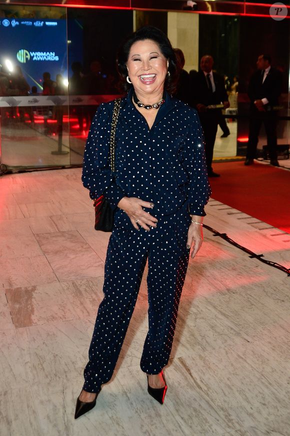 Dona Kika veste um conjunto azul-marinho com estampa de pequenas bolinhas claras, composto por camisa de mangas longas e calça de corte reto, criando um visual sofisticado com toque retrô. O look é arrematado com bolsa preta de corrente dourada e scarpins escuros