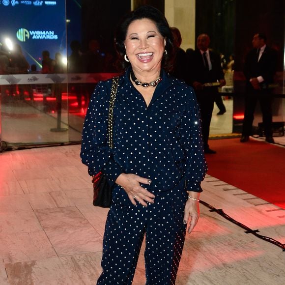 Dona Kika veste um conjunto azul-marinho com estampa de pequenas bolinhas claras, composto por camisa de mangas longas e calça de corte reto, criando um visual sofisticado com toque retrô. O look é arrematado com bolsa preta de corrente dourada e scarpins escuros