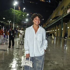 Duda Lopes investiu em uma proposta oversized composta por camisão branco de botões com mangas compridas dobradas no pulso e calça jeans bem ampla estilo pantalona com lavagem manchada/estonada. Acessórios neutros complementaram o visual, incluindo uma bolsa tiracolo estruturada de formato geométrico em tom off-white, colar com pingente de chave, anéis e chinelos de dedo pretos