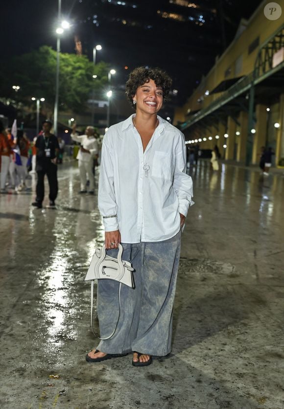 Duda Lopes investiu em uma proposta oversized composta por camisão branco de botões com mangas compridas dobradas no pulso e calça jeans bem ampla estilo pantalona com lavagem manchada/estonada. Acessórios neutros complementaram o visual, incluindo uma bolsa tiracolo estruturada de formato geométrico em tom off-white, colar com pingente de chave, anéis e chinelos de dedo pretos