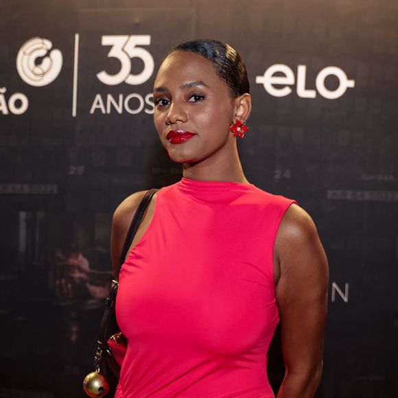 No tapete vermelho, Jéssica Ellen aposta em um visual monocromático de forte impacto, com vestido midi em tom vibrante de vermelho que valoriza a silhueta por meio de drapeados estratégicos ao longo do corpo, criando textura e movimento sutil; a peça, de gola alta e sem mangas, reforça a elegância minimalista, enquanto os acessórios seguem a mesma cartela, com sandálias de tiras finas e brilho acetinado e brincos esculturais que conversam com o look, além de uma bolsa de alça fina com detalhe metálico