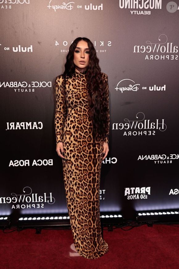 Sabrina Viana veste um vestido longo e justo ao corpo com gola alta e mangas compridas, confeccionado inteiramente em padronagem de animal print de onça clássica. O visual clean e elegante é finalizado com cabelos bem longos e ondulados soltos divididos ao meio, maquiagem suave com destaque nos olhos e unhas compridas escuras