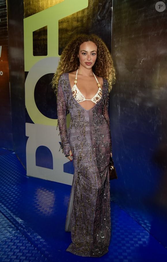 Julia Rodrigues surgiu com uma proposta que exala sofisticação e transparência. O look consiste em um vestido longo de mangas compridas em tecido fluido com estampa abstrata em tons de lavanda e cinza, ricamente adornado com bordados curvilíneos em linha e pedraria escura. O diferencial fica por conta do decote profundo que deixa à mostra um top estilo biquíni em tom off-white, com bordados étnicos em marrom e búzios aplicados nas alças