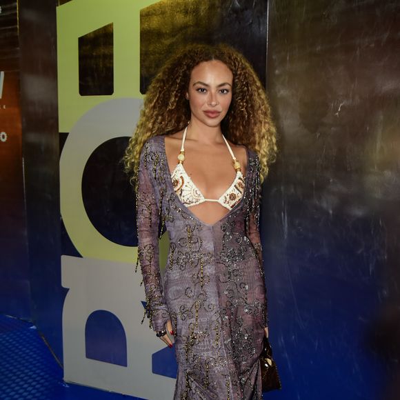 Julia Rodrigues surgiu com uma proposta que exala sofisticação e transparência. O look consiste em um vestido longo de mangas compridas em tecido fluido com estampa abstrata em tons de lavanda e cinza, ricamente adornado com bordados curvilíneos em linha e pedraria escura. O diferencial fica por conta do decote profundo que deixa à mostra um top estilo biquíni em tom off-white, com bordados étnicos em marrom e búzios aplicados nas alças