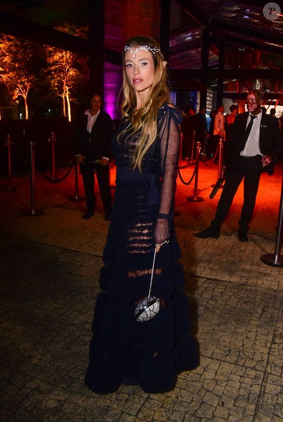 Juliana Didone veste um vestido longo preto de camadas sobrepostas com babados transparentes e mangas de tule pretas bufantes com amarrações nos ombros. Ela complementa a produção com uma tiara de elfo prateada com strass na testa, maquiagem com brilho nos olhos e uma pequena bolsa esférica metálica prateada com alça de corrente. O visual tem uma temática de elfo
