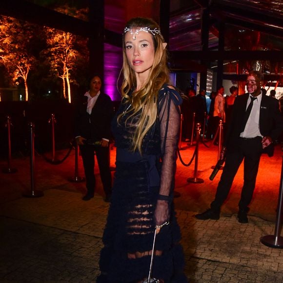 Juliana Didone veste um vestido longo preto de camadas sobrepostas com babados transparentes e mangas de tule pretas bufantes com amarrações nos ombros. Ela complementa a produção com uma tiara de elfo prateada com strass na testa, maquiagem com brilho nos olhos e uma pequena bolsa esférica metálica prateada com alça de corrente. O visual tem uma temática de elfo