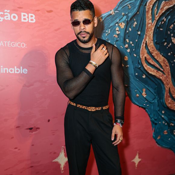 Felipe Velozo investiu na tendência da transparência com uma blusa de tela preta ajustada ao corpo, destacando os braços. A peça foi combinada com uma calça de alfaiataria preta de cintura alta, marcada por um cinto fino com estampa de leopardo (animal print) que quebra a sobriedade do look total black. Óculos de sol de lentes escuras e retangulares, junto a sapatos de sola tratorada, conferem um ar urbano e rocker à produção