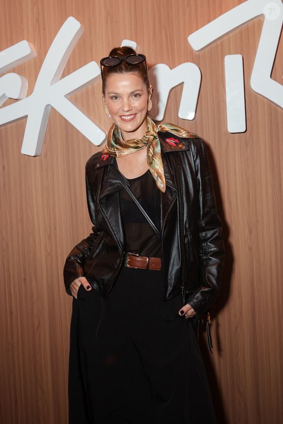 Para este evento, a influenciadora escolheu um look sofisticado e com um toque moderno. Ela veste uma jaqueta de couro preta estruturada, com detalhes de zíper e lapela, sobreposta a uma blusa de tule transparente e um top preto por baixo, o que cria um jogo de texturas interessante. Na cintura, um cinto de couro marrom com fivela dourada define a silhueta. Nos acessórios, o destaque fica para o lenço de seda com estampa floral e tons de verde e dourado amarrado no pescoço, que quebra o visual monocromático e adiciona um toque de cor e feminilidade