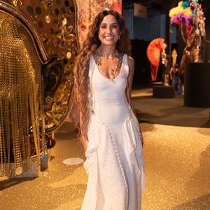 Camila Pitanga iluminou o evento com uma estética 'boho-chic' solar e sofisticada, vestindo um longo da grife Aluf em tom off-white. O vestido, confeccionado em um tecido de textura rústica que remete ao linho tramado, apresenta um decote em 'V' profundo e uma modelagem escultural com recortes verticais que terminam em barras levemente desfiadas, conferindo um ar artesanal de luxo