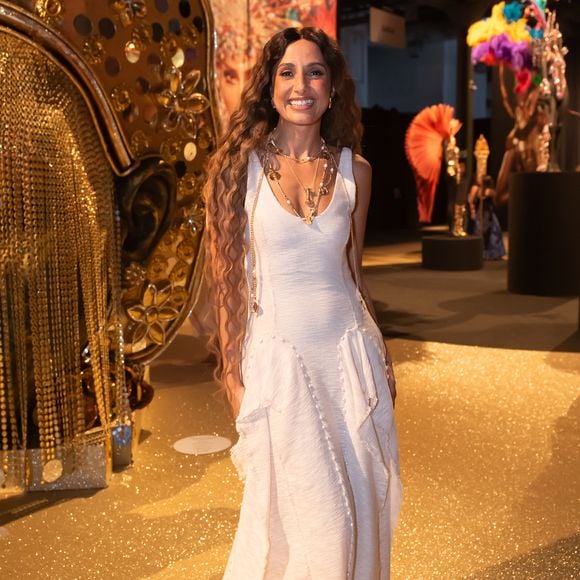 Camila Pitanga iluminou o evento com uma estética 'boho-chic' solar e sofisticada, vestindo um longo da grife Aluf em tom off-white. O vestido, confeccionado em um tecido de textura rústica que remete ao linho tramado, apresenta um decote em 'V' profundo e uma modelagem escultural com recortes verticais que terminam em barras levemente desfiadas, conferindo um ar artesanal de luxo