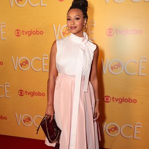 Sheron Menezzes, protagonista de 'Por Você', apostou em um look clássico e feminino com um toque contemporâneo. Ela veste uma blusa branca de seda com gola alta e laço no pescoço, combinada com uma saia rodada rosa claro com fenda frontal. Para completar o visual, escolheu sapatos scarpin dourados metalizados e uma bolsa de mão em marrom escuro. Seus cabelos estão presos em um coque alto e trançado