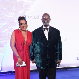 Péricles e Lidiane: ela em coque alto usa vestido longo vermelho de decote profundo com detalhes em ferragens prateadas no busto, complementado por bolsa clutch dourada. Ele, de óculos e barba grisalha, veste terno com paletó de veludo verde-escuro, lapela preta texturizada brilhante, camisa branca e gravata borboleta preta