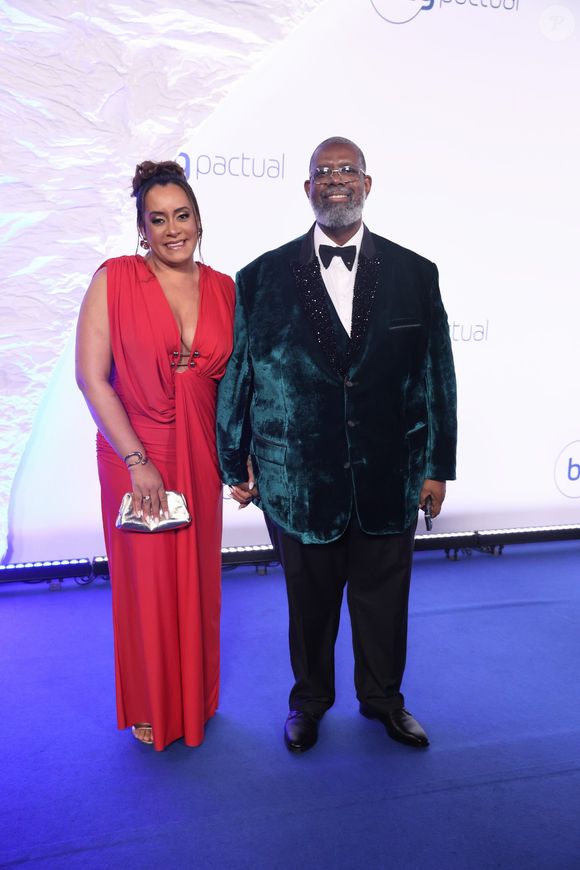 Péricles e Lidiane: ela em coque alto usa vestido longo vermelho de decote profundo com detalhes em ferragens prateadas no busto, complementado por bolsa clutch dourada. Ele, de óculos e barba grisalha, veste terno com paletó de veludo verde-escuro, lapela preta texturizada brilhante, camisa branca e gravata borboleta preta