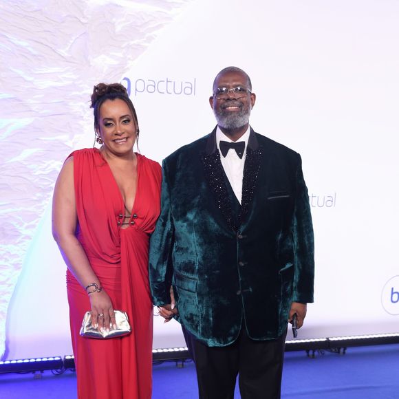 Péricles e Lidiane: ela em coque alto usa vestido longo vermelho de decote profundo com detalhes em ferragens prateadas no busto, complementado por bolsa clutch dourada. Ele, de óculos e barba grisalha, veste terno com paletó de veludo verde-escuro, lapela preta texturizada brilhante, camisa branca e gravata borboleta preta