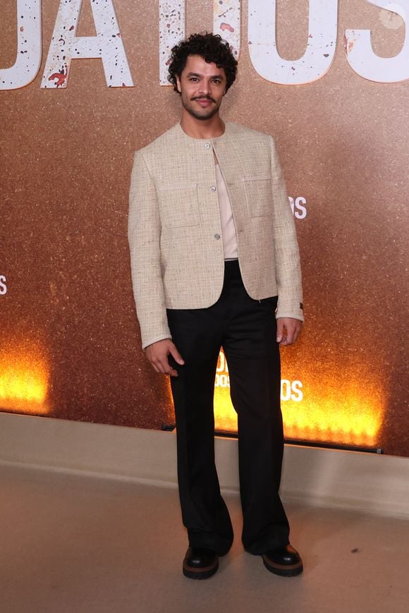 Matheus Abreu veste um casaco curto de tweed bege sem gola, com abotoamento frontal e bolsos aplicados, sobreposto a uma camisa neutra de gola simples. A produção equilibra o clássico com o urbano ao combinar calça preta de alfaiataria reta a sapatos tratorados de sola robusta