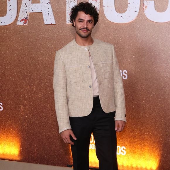 Matheus Abreu veste um casaco curto de tweed bege sem gola, com abotoamento frontal e bolsos aplicados, sobreposto a uma camisa neutra de gola simples. A produção equilibra o clássico com o urbano ao combinar calça preta de alfaiataria reta a sapatos tratorados de sola robusta
