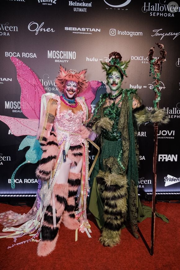 Fih e Edu, do 'Divas Depressão': o casal surge com fantasias coordenadas de inspiração na floresta e no universo felino; Fih usa um corpete rosa com brilhantes, asas de borboleta em tom rosa e turquesa, detalhes em pelagem estampada de tigre e cartola com orelhas. Edu veste uma túnica drapeada em tons de verde com texturas folhosas, perneiras felpudas de pelagem verde e preta, maquiagem felina e empunha um cajado de madeira esculpido com folhagens enroladas